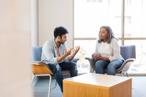 The young adult man gestures emphatically as he shares with the serious senior adult female therapist.