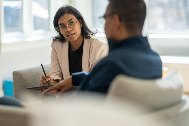 A middle aged gentleman sits across from his female therapist as they talk through his struggles. The Therapist is dressed professionally and taking notes on a tablet.